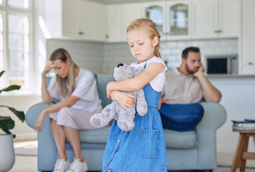 A girl clutches a teddy bear, her expression wistful, as a woman and man sit on a sofa in the background, their faces etched with the distress of pondering how long can a divorce be put on hold in the tense stillness of the living room.