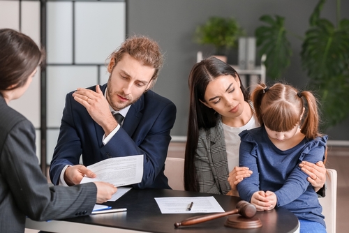 Two adults and a child sit at a table with legal documents and a gavel. One adult points at a paper, possibly discussing how long a divorce can be put on hold, while the other comforts the child. A person across the table listens intently.