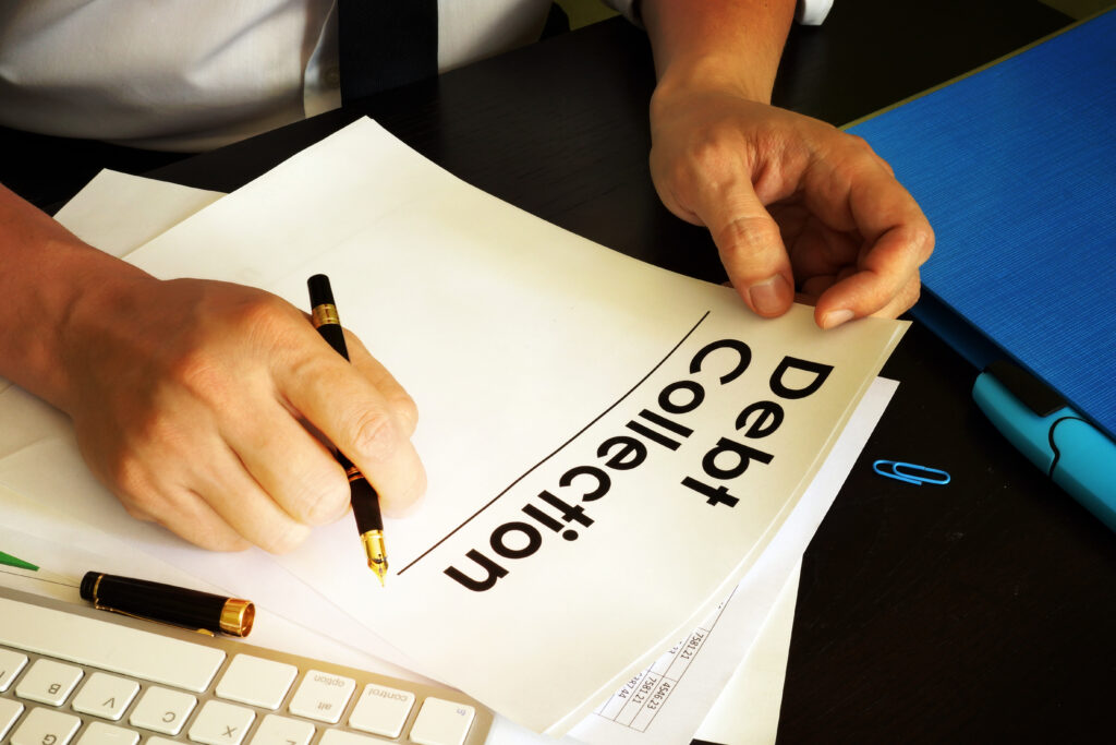 A person holding a document labeled "Debt Collection" and preparing to sign it with a pen. A keyboard, a calculator, and other office supplies are visible on the desk.