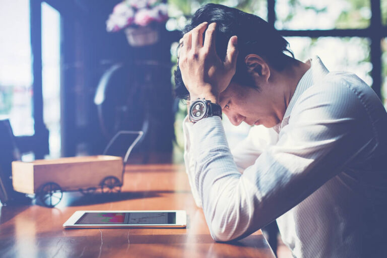 A man sitting at a desk with a tablet sitting flat on the table and the mans elbows are resting on the table with his head in his hands