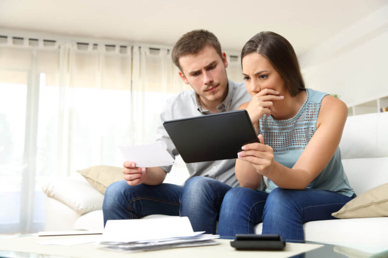 Man and woman sitting on the couch, the woman is holding a tablet they both are looking at and sitting in front of them on the coffee table is a stack of papers