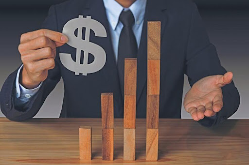 A person in a suit holds a dollar sign next to an increasing bar graph made of wooden blocks, symbolizing financial growth.