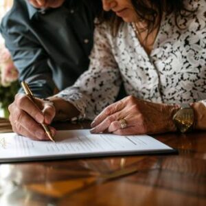 Two people signing a prenup at a wooden table, with one person holding the pen and the other guiding or observing, making sure their agreement is enforceable under Texas law.