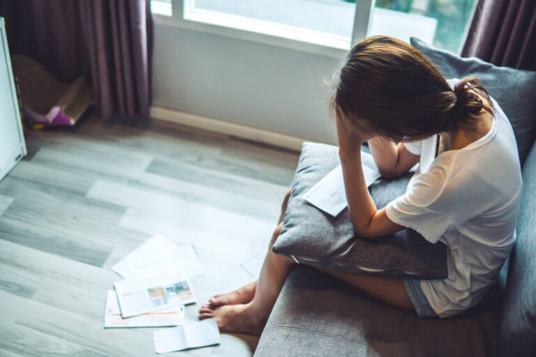 A woman sitting on the couch with a pillow on her lap and a piece of paper on top of the pillow, on the ground there are many different papers spread out