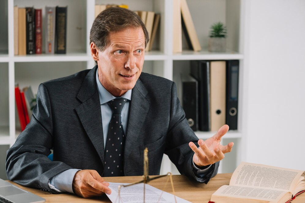 A man in a suit sits in an office, holding documents and gesturing with one hand while discussing the pros and cons of declaring bankruptcy, with a bookshelf in the background.