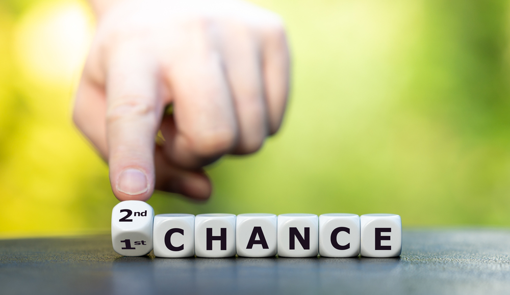 Hand rearranging dice spelling "CHANCE" to display "SECOND CHANCE" on a blurred green background, inviting viewers to weigh their options.