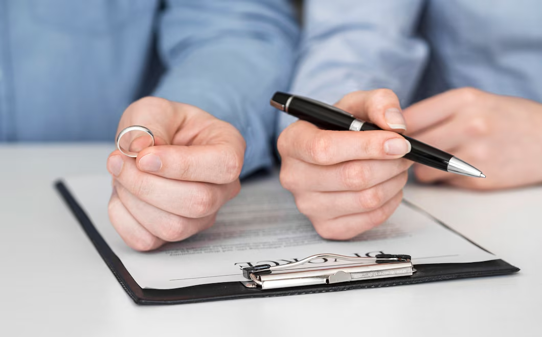 Two people in blue shirts sit at a desk holding a wedding ring and a pen over a document with the word "DIVORCE" visible. 