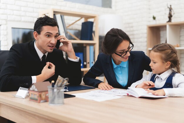 Two professionals in business attire discussing documents with a young girl at a desk.