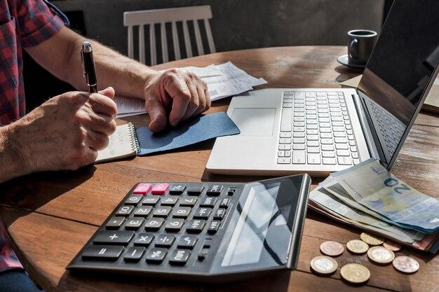 A person making financial calculations with a calculator, notepad, and laptop amidst scattered money.