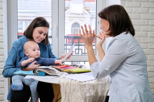 Woman with a baby in her lap consulting with another woman across a table with fabric samples.