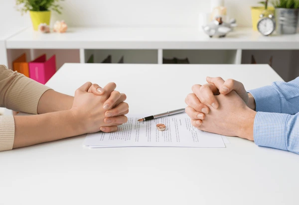 Two people sitting at a table with clasped hands, with a divorce document and pen between them.