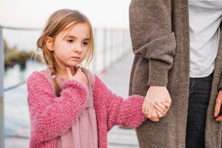 A young girl in a pink sweater holds an adult's hand, looking thoughtful, on a bridge at dusk.