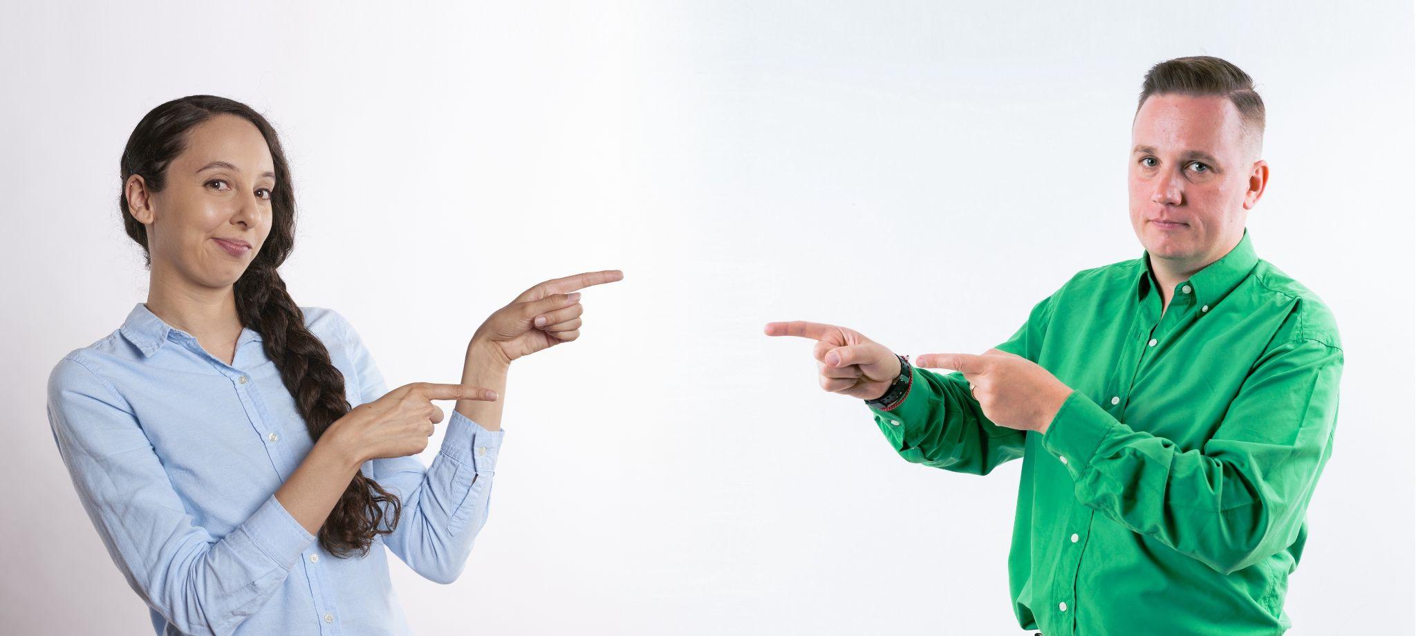 A man and woman are pointing at each other. The woman, with long dark hair in a light blue shirt, looks stern. The man, sporting short hair and a green shirt, has an equally serious expression. 