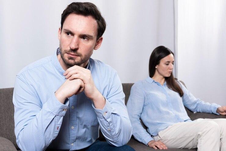 Two individuals in light blue shirts sit apart on a couch, both looking away from each other with serious expressions
