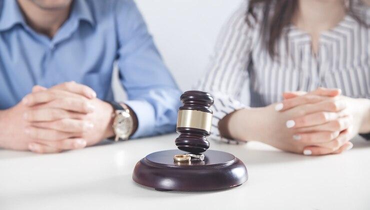 Two individuals seated with hands clasped on a table, a judge's gavel on a stand between them, and a pair of wedding rings near the gavel. 