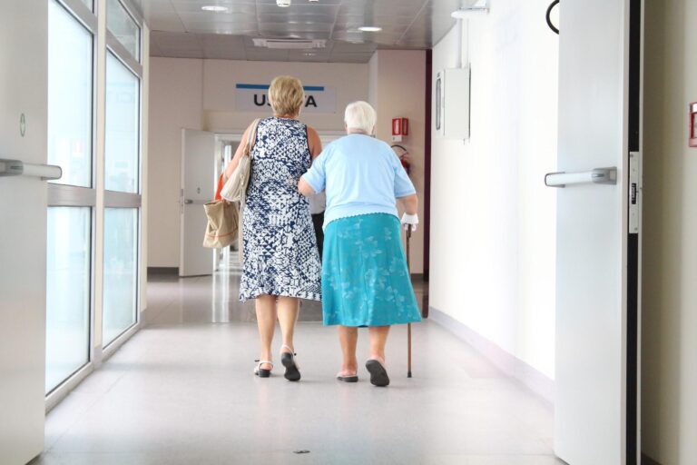 Two elderly women, one using a cane, walk down a well-lit corridor towards a door marked "U.R.A" in a medical facility.