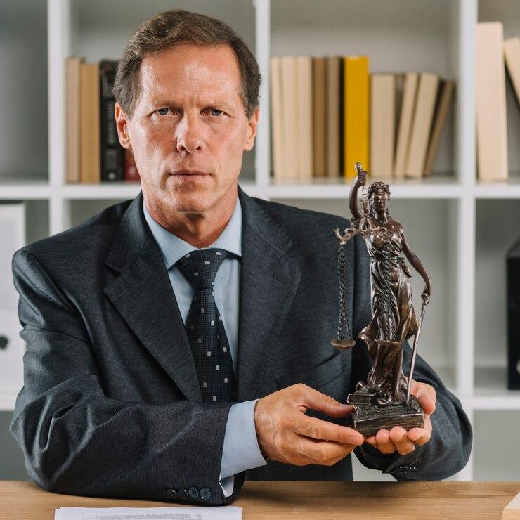 A man in a suit holds a small Lady Justice statue while sitting at a desk with shelves of books in the background.