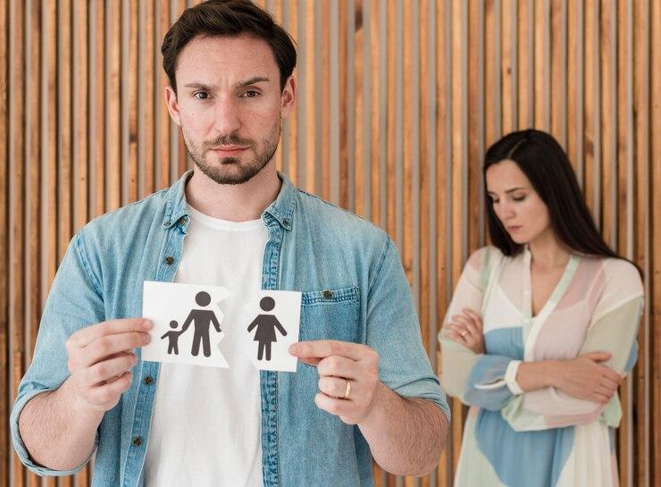 A man holds a torn paper showing a family icon, while a woman stands in the background with folded arms, both looking distressed.