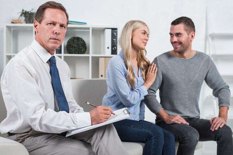 A man in a white shirt takes notes while sitting on a couch. A woman and a man sit together, with the woman placing her hand on the man's shoulder.