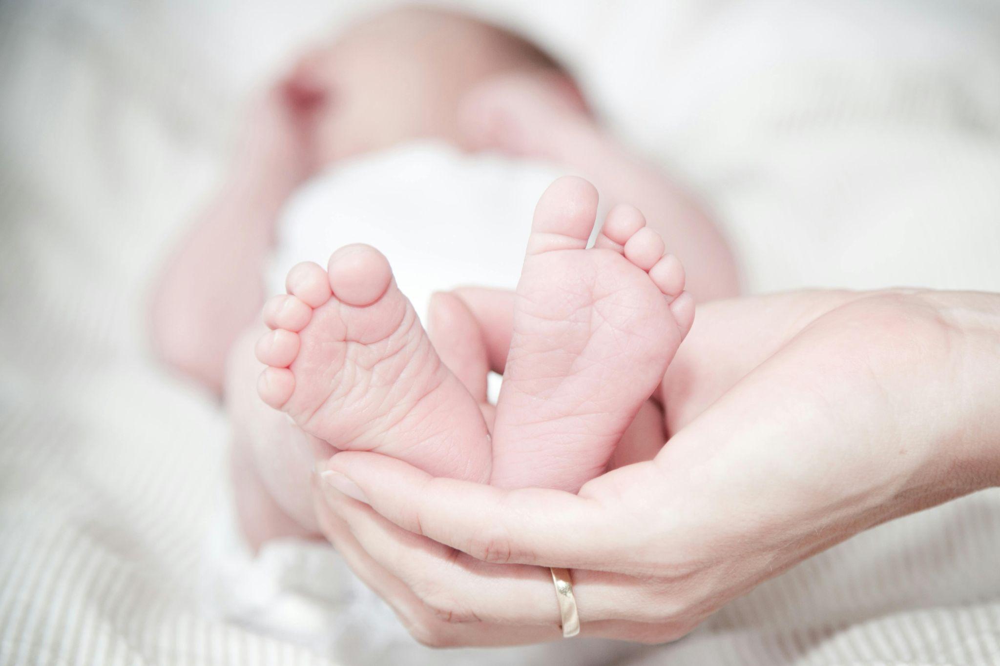 A person gently holds a baby's feet in their hand, with a gold ring visible on their finger, symbolizing hope even as parents explore compensation options for birth-related injuries. The baby's face is out of focus in the background.