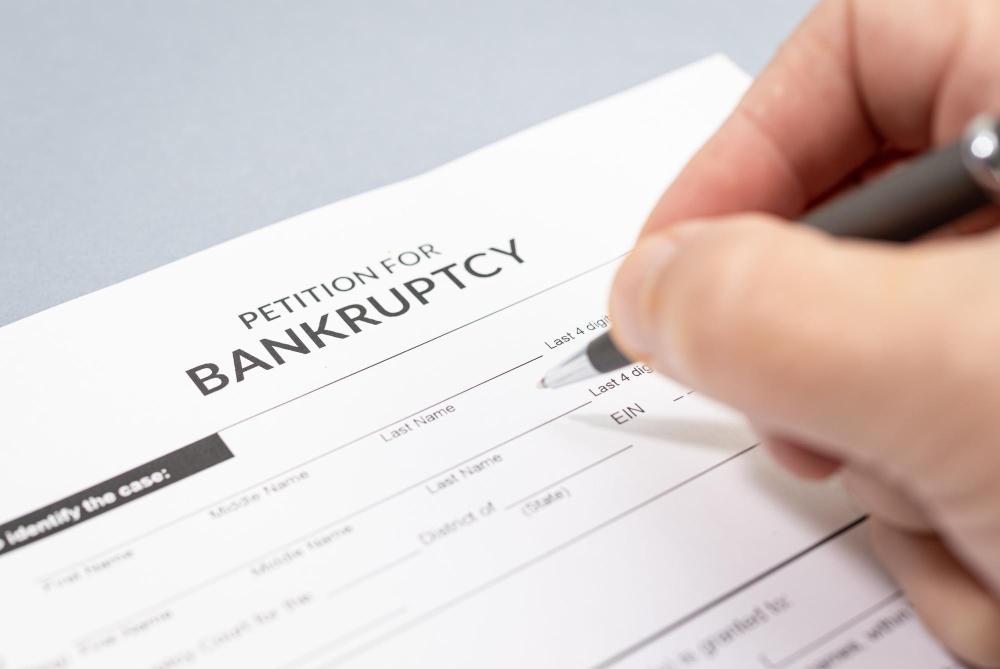 A person holds a pen to fill out a "Petition for Bankruptcy" form with blank spaces for personal and financial information. The form includes fields for a petitioner's name, details, and case number.