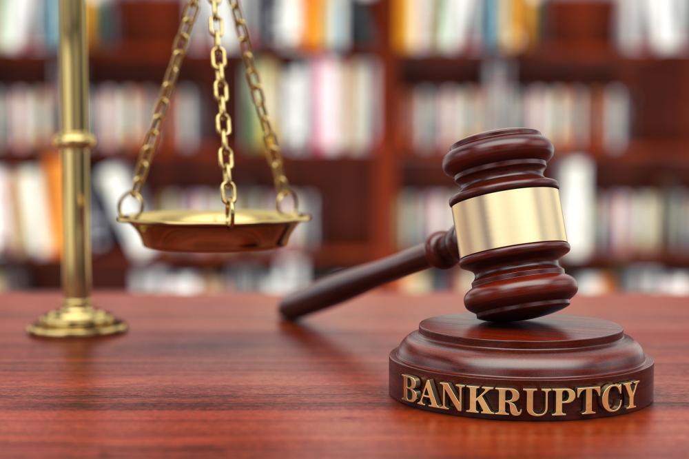 A gavel labeled "Bankruptcy" lies on a wooden surface beside a blurred background featuring a scale of justice and a bookshelf.