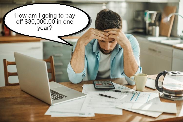 A man sits at a kitchen table with papers, a calculator, and a laptop, looking stressed. A thought bubble above reads, "How am I going to pay off $30,000.00 in one year on a low income?