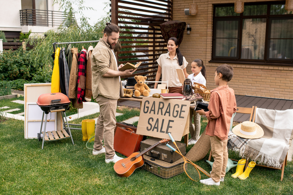 A family participates in a garage sale outside their home, displaying various items like clothes, a guitar, toys, and home decor on tables and racks—an excellent way to learn how to get out of debt on a low income.