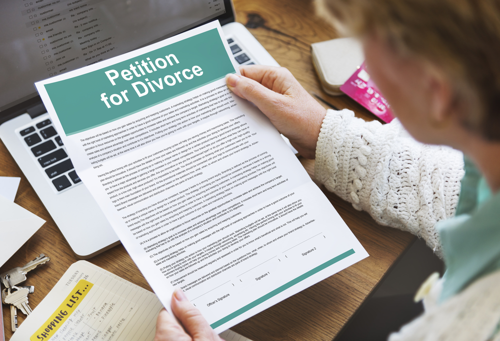 A person sits at a table, anxiously pondering the implications of divorce, as they glance at a petition for divorce document beside their laptop, notebook, and pen.